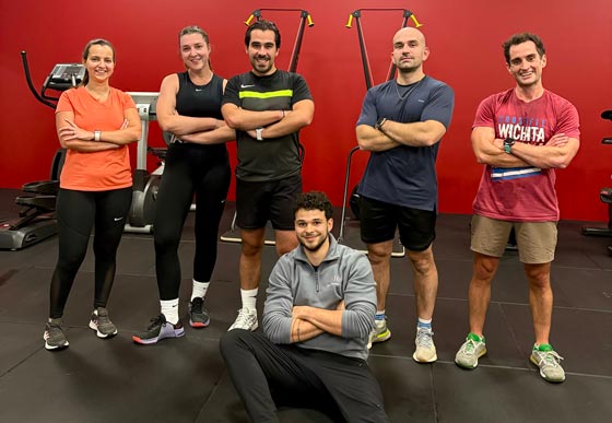 Une équipe de six sportifs pose fièrement bras croisés dans une salle de fitness aux murs rouges.
