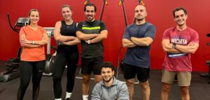 Une équipe de six sportifs pose fièrement bras croisés dans une salle de fitness aux murs rouges.