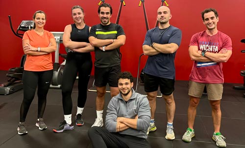 Une équipe de six sportifs pose fièrement bras croisés dans une salle de fitness aux murs rouges.