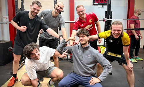 Un groupe d'hommes souriants partage un moment de convivialité dans une salle de musculation équipée de racks.