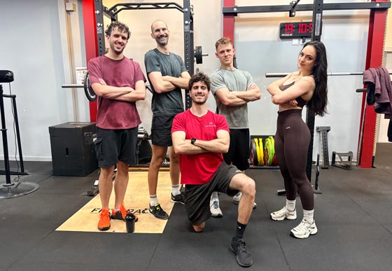 Quatre hommes et une femme posent avec assurance devant une cage de squat dans une salle de sport moderne.
