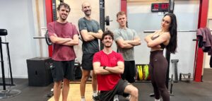 Quatre hommes et une femme posent avec assurance devant une cage de squat dans une salle de sport moderne.