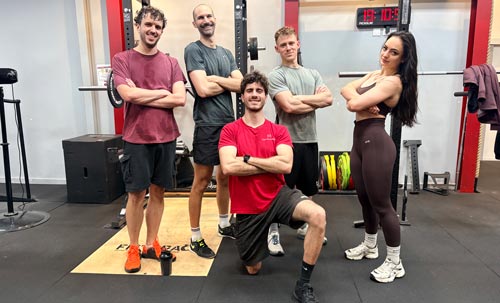 Quatre hommes et une femme posent avec assurance devant une cage de squat dans une salle de sport moderne.